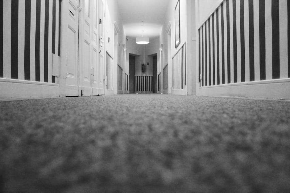 A person using a yellow vacuum cleaner with a black hose attachment to perform deep cleaning on a patterned residential carpet in a room with soft lighting. The individual is dressed casually in a bei