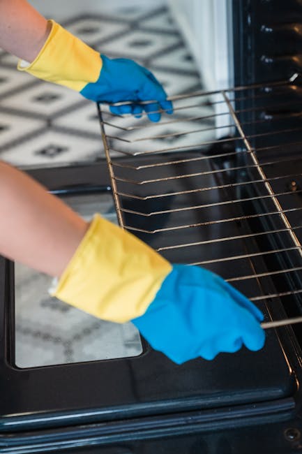 Close-up of a person wearing yellow and blue rubber gloves, using their hands to handle a wire rack inside an oven on Lonsdale Road Queens Park. The oven interior appears clean and free of food debris